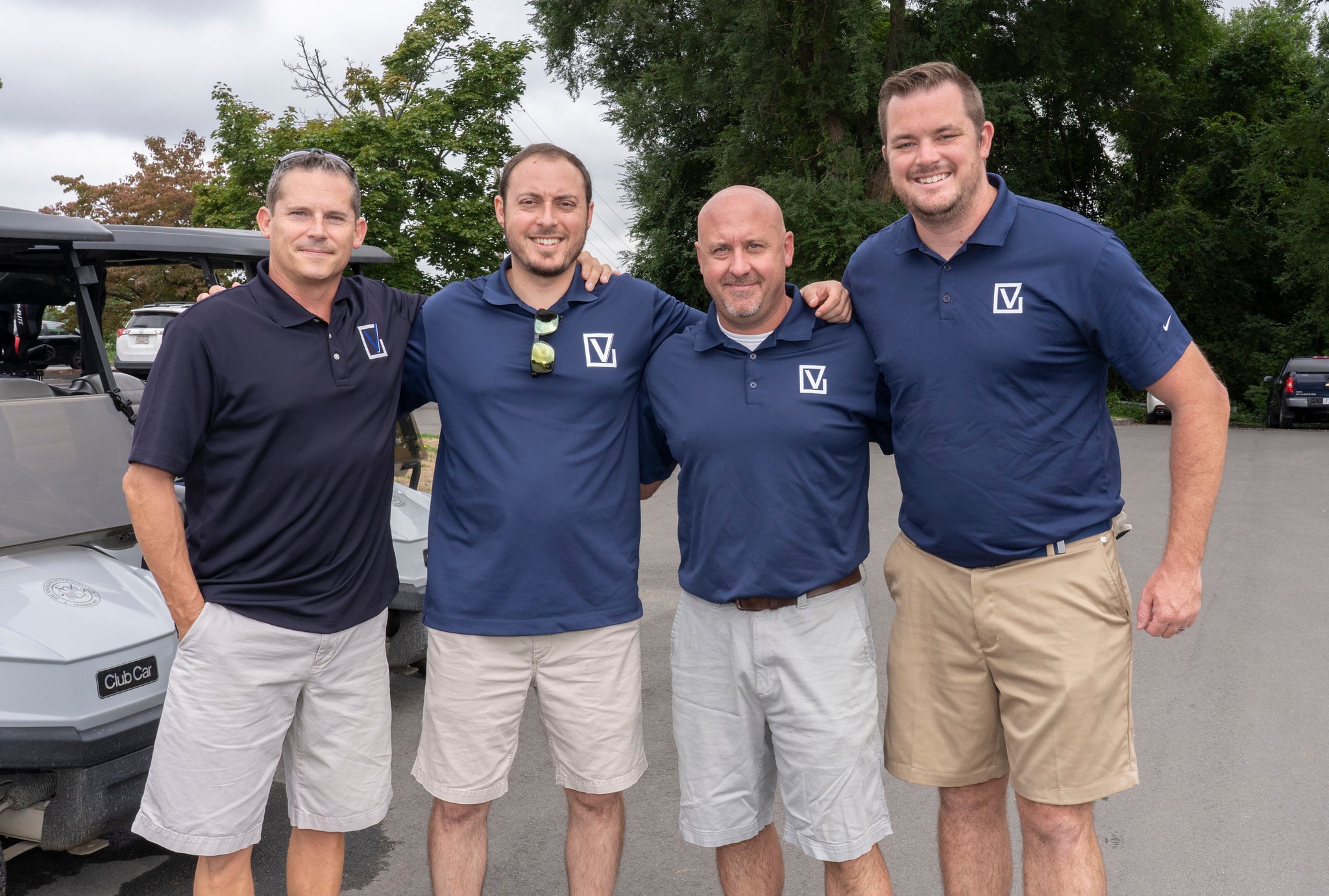 Four men wearing matching navy tees and khaki shorts, ready to get in their golf carts and play.